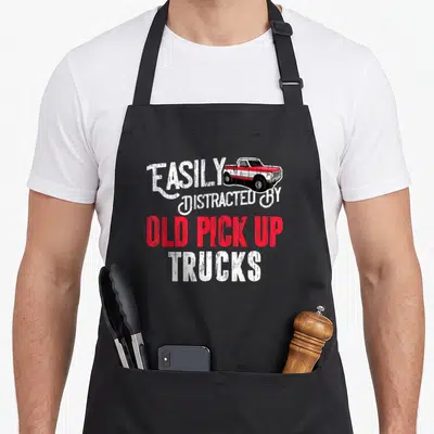 Distracted pickup truck apron - Distressed white and red text reading EASILY DISTRACTED BY OLD PICK UP TRUCKS — shown on a black cotton canvas apron against a white background