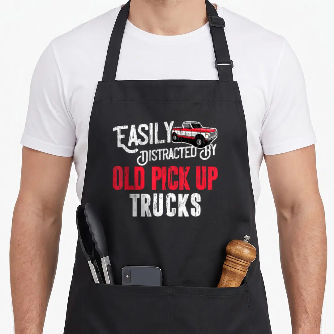 Distracted pickup truck apron - Distressed white and red text reading EASILY DISTRACTED BY OLD PICK UP TRUCKS — shown on a black cotton canvas apron against a white background