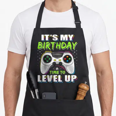 Vintage level video game apron - Cartoon controller surrounded by stars with IT'S MY BIRTHDAY TIME TO LEVEL UP text — shown on a black cotton canvas apron against a white background