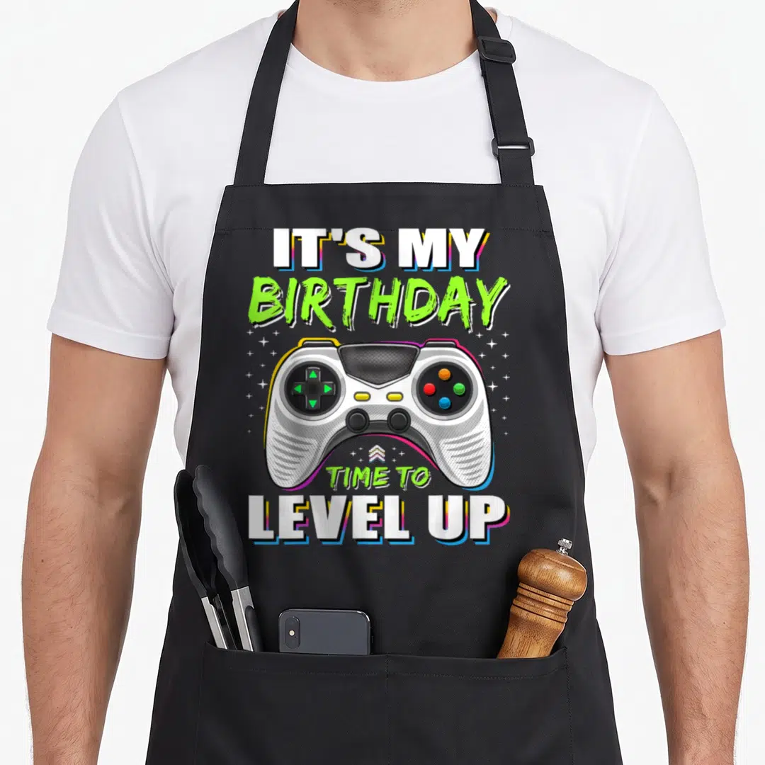 Vintage level video game apron - Cartoon controller surrounded by stars with IT'S MY BIRTHDAY TIME TO LEVEL UP text — shown on a black cotton canvas apron against a white background