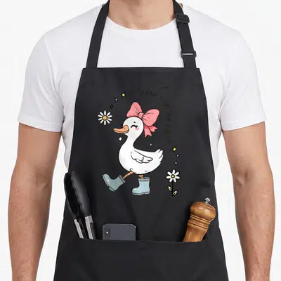 Retro goose silly apron - Cartoon white goose with pink bow and blue boots under arched text surrounded by flowers — shown on a black cotton canvas apron against a white background