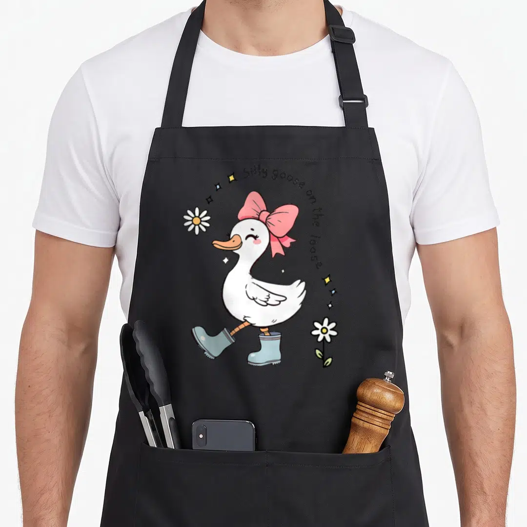 Retro goose silly apron - Cartoon white goose with pink bow and blue boots under arched text surrounded by flowers — shown on a black cotton canvas apron against a white background