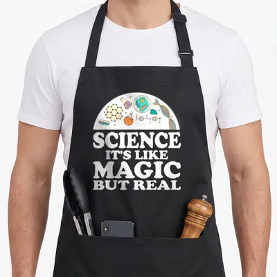 Magic but science apron - White semi-circle with cartoon lab icons above stacked text SCIENCE IT'S LIKE MAGIC BUT REAL — shown on a black cotton canvas apron against a white background