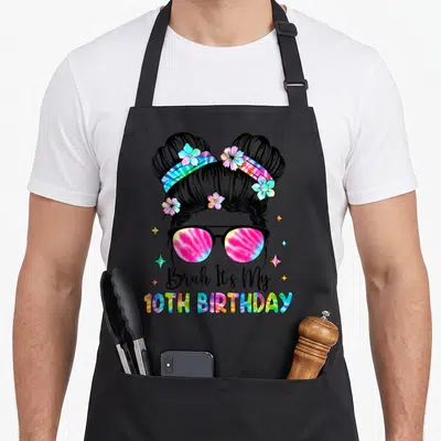 Bruh 10th birthday apron - Cartoon girl with space buns and tie-dye sunglasses holding Bruh It's My 10TH BIRTHDAY text — shown on a black cotton canvas apron against a white background