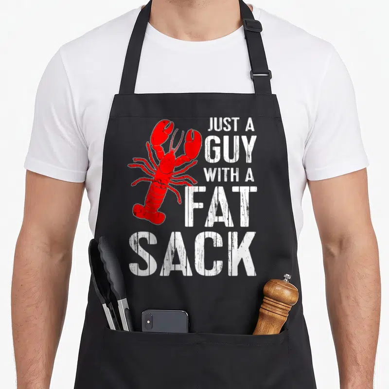 Crawfish fat man in apron - Red lobster illustration next to stacked white distressed text reading JUST A GUY — shown on a black cotton canvas apron against a white background