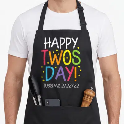 Twosday teacher apron - HAPPY TWOS DAY! text in pink orange yellow green blue purple with small yellow stars — shown on a black cotton canvas apron against a white background