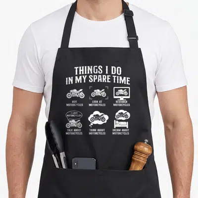 Spare biker apron - White line art of motorcycles with text listing spare time activities on black fabric — shown on a black cotton canvas apron against a white background