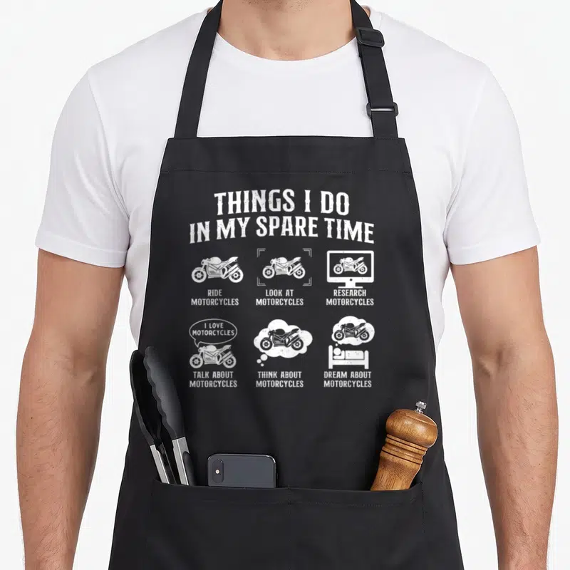 Spare biker apron - White line art of motorcycles with text listing spare time activities on black fabric — shown on a black cotton canvas apron against a white background