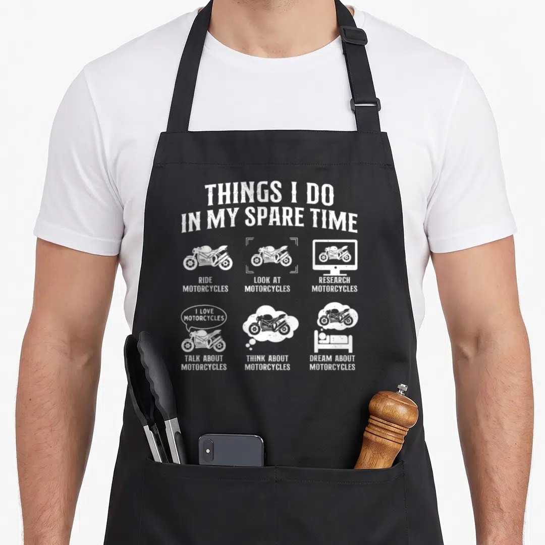 Spare biker apron - White line art of motorcycles with text listing spare time activities on black fabric — shown on a black cotton canvas apron against a white background