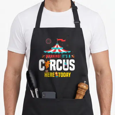 Zirkus birthday apron - Striped circus tent with WARNING text, cartoon bear, lion, monkeys, and fireworks in red white — shown on a black cotton canvas apron against a white background