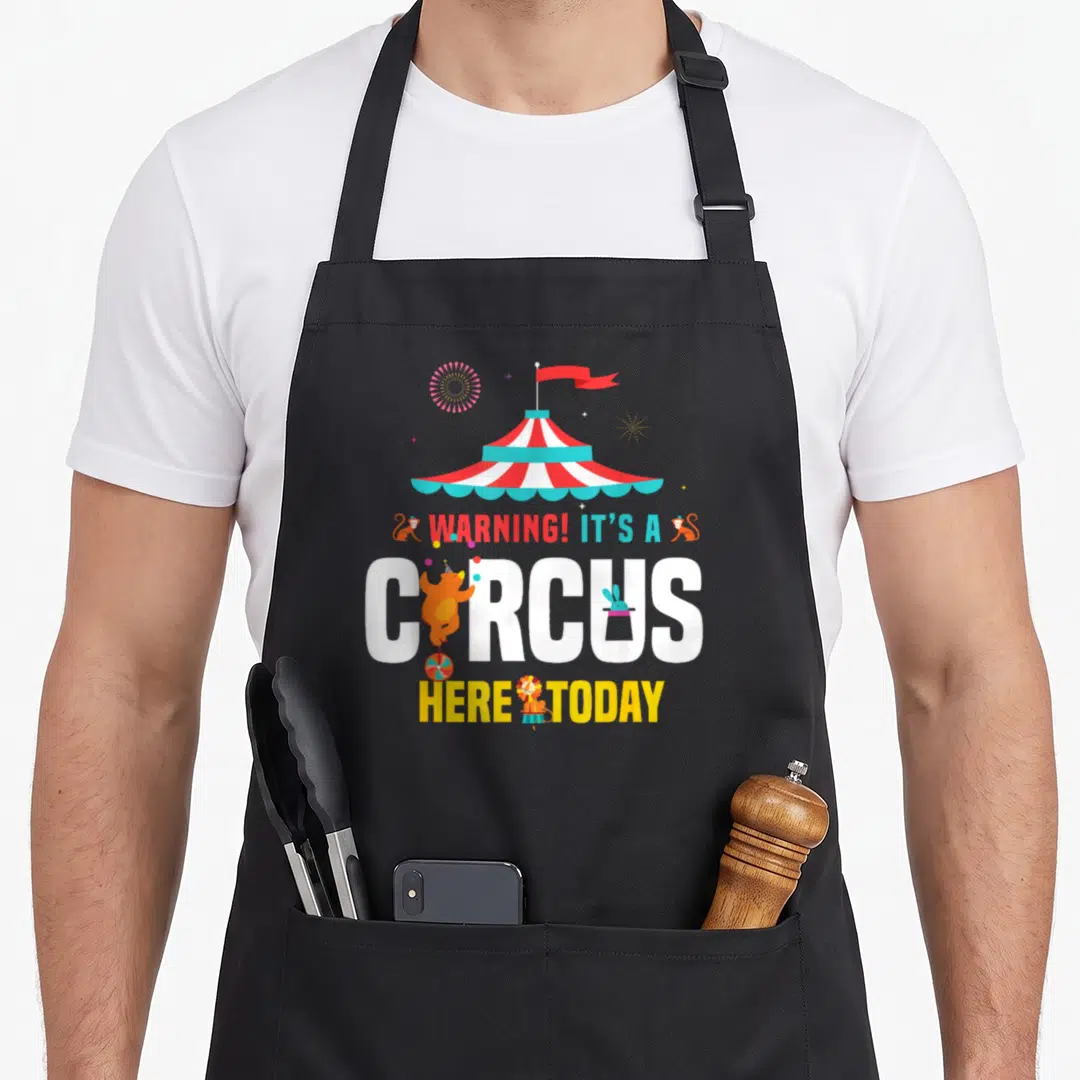 Zirkus birthday apron - Striped circus tent with WARNING text, cartoon bear, lion, monkeys, and fireworks in red white — shown on a black cotton canvas apron against a white background