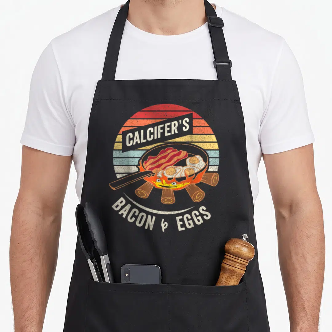 Calcifer bacon apron - Retro frying pan with bacon eggs over campfire and striped sun background — shown on a black cotton canvas apron against a white background