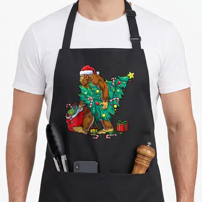 Most christmas tree apron - Cartoon Bigfoot carrying green Christmas tree with red sack and scattered candy canes — shown on a black cotton canvas apron against a white background