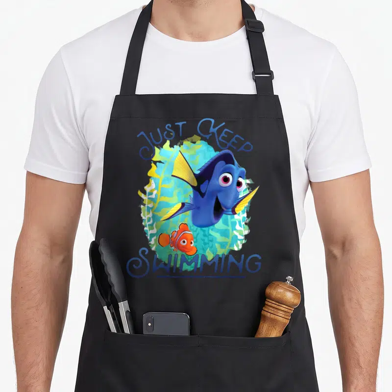 Dory swimming apron - Blue and orange fish with Just Keep Swimming text over green seaweed — shown on a black cotton canvas apron against a white background