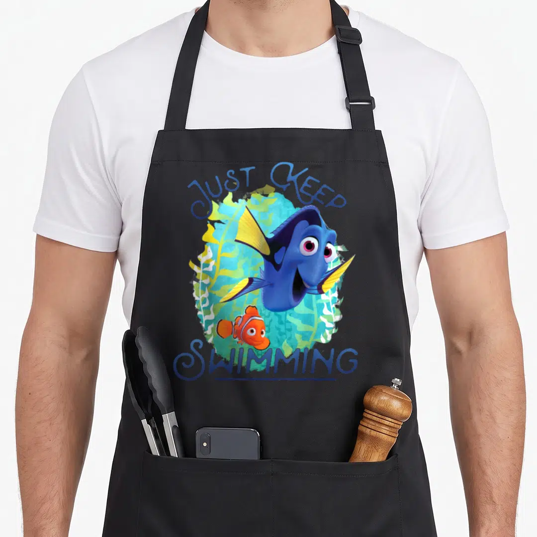 Dory swimming apron - Blue and orange fish with Just Keep Swimming text over green seaweed — shown on a black cotton canvas apron against a white background
