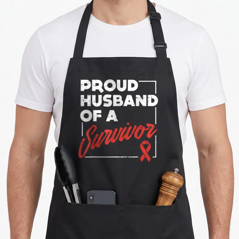 Stroke husband apron - Vertical white text reading PROUD HUSBAND OF A Survivor with red ribbon in corner — shown on a black cotton canvas apron against a white background