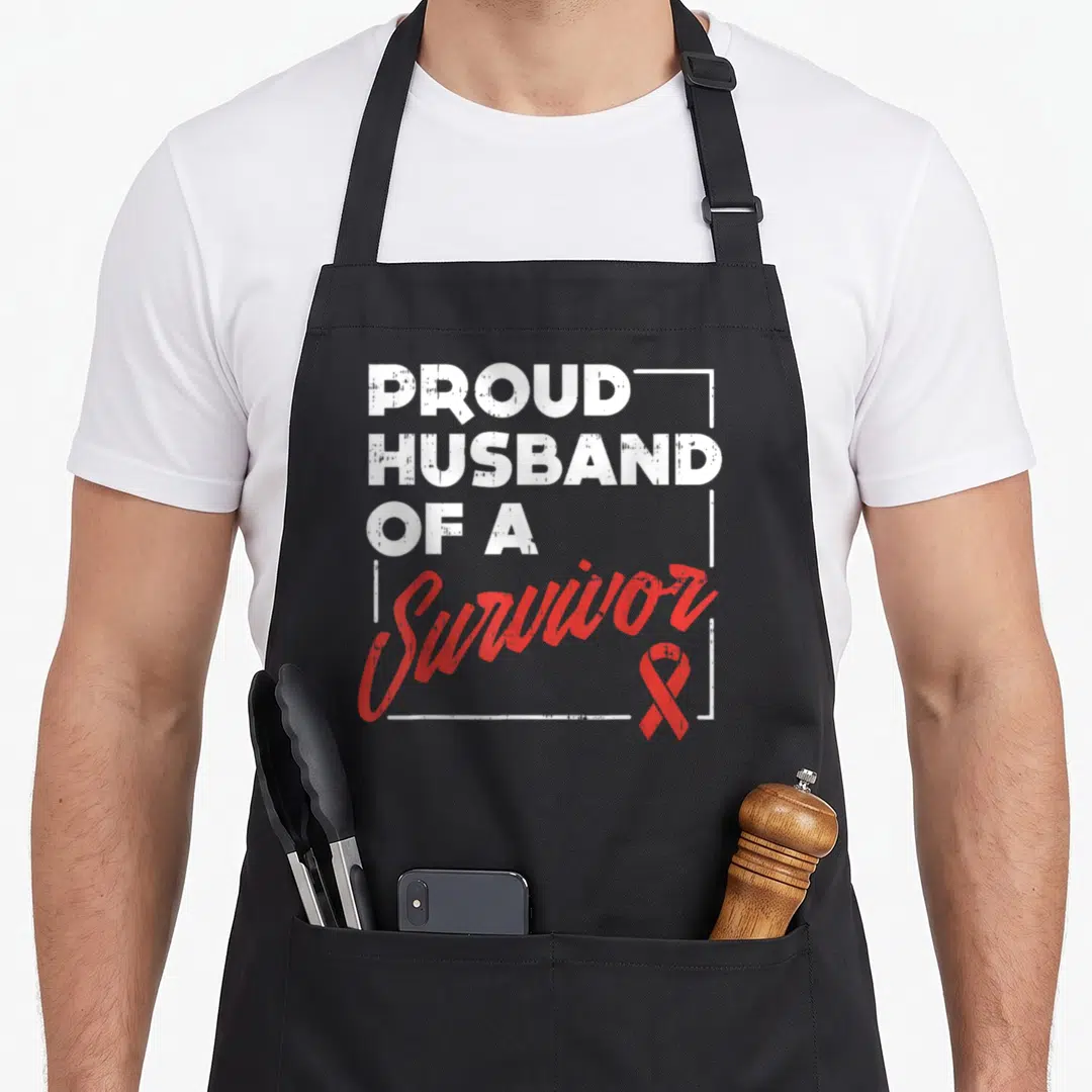 Stroke husband apron - Vertical white text reading PROUD HUSBAND OF A Survivor with red ribbon in corner — shown on a black cotton canvas apron against a white background