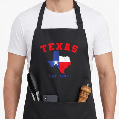 Est texans apron - Arched TEXAS text above Texas map filled with red white blue flag and EST 1845 below — shown on a black cotton canvas apron against a white background