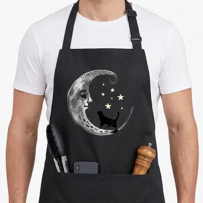 Star black cat apron - Crescent moon with face, black cat silhouette, and yellow stars illustration — shown on a black cotton canvas apron against a white background