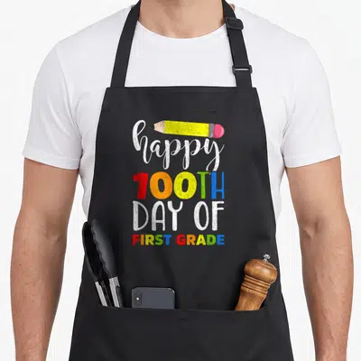 100th grade teacher apron - Happy 100th Day of First Grade text with yellow pencil and multi-colored letters — shown on a black cotton canvas apron against a white background