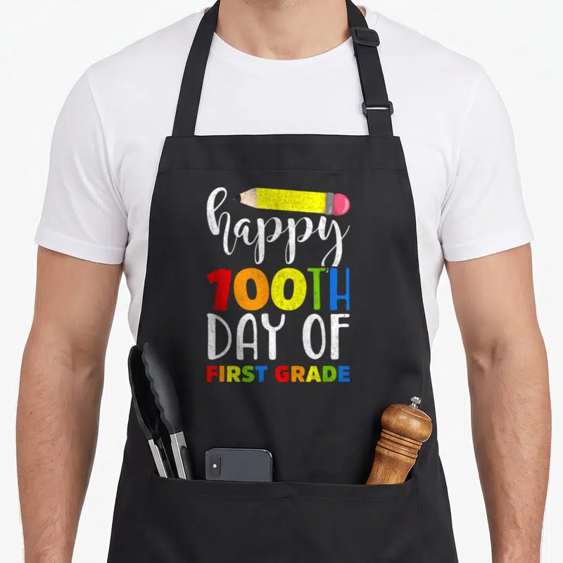 100th grade teacher apron - Happy 100th Day of First Grade text with yellow pencil and multi-colored letters — shown on a black cotton canvas apron against a white background