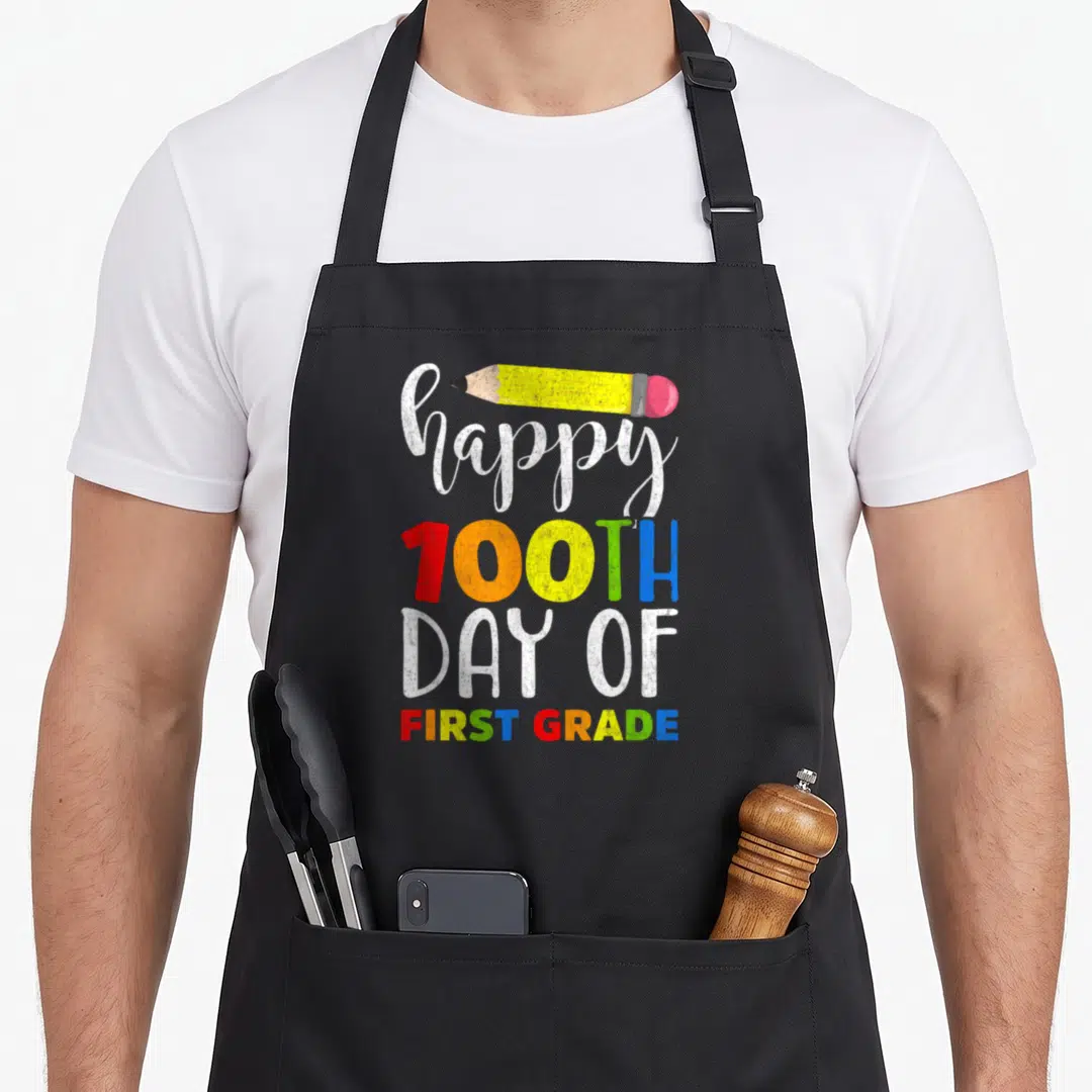 100th grade teacher apron - Happy 100th Day of First Grade text with yellow pencil and multi-colored letters — shown on a black cotton canvas apron against a white background