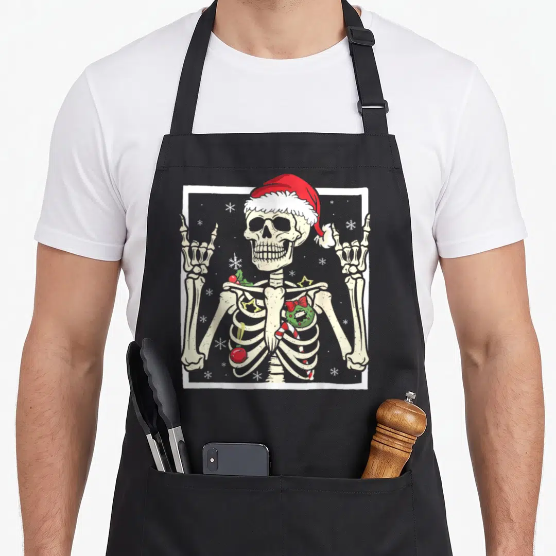 Santa punk rock apron - Cartoon skeleton in red hat with holly and candy cane ornaments in ribcage surrounded — shown on a black cotton canvas apron against a white background