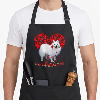 Eskimo dog apron - White fluffy dog holding red rose in front of red paw print heart — shown on a black cotton canvas apron against a white background