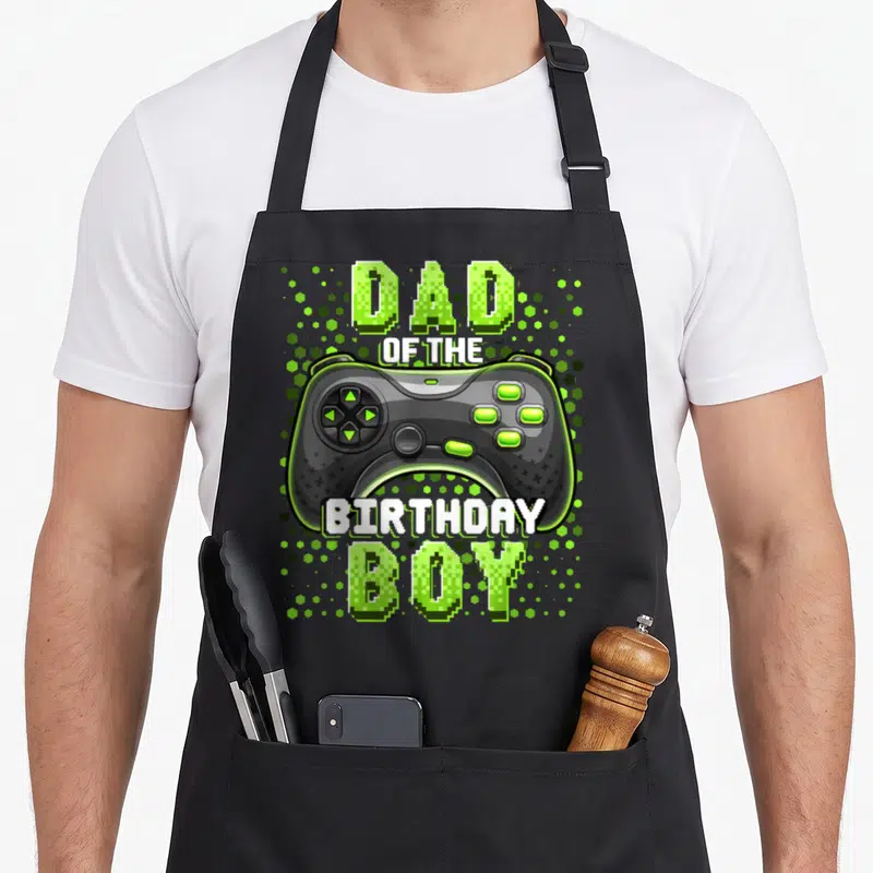 Cool video birthday apron - Game controller graphic with Dad of the Birthday Boy text and green hexagons — shown on a black cotton canvas apron against a white background