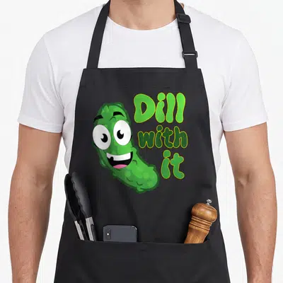Cute dill pickle apron - Cartoon pickle character next to green Dill with it text — shown on a black cotton canvas apron against a white background
