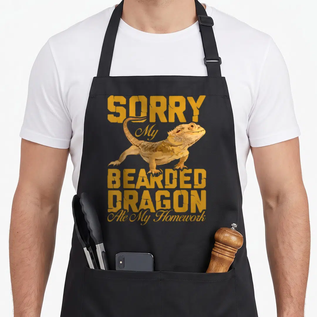 Homework dragon apron - Yellow and tan bearded dragon illustration with sorry ate homework text — shown on a black cotton canvas apron against a white background