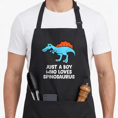Retro spinosaurus dinosaur apron - Blue cartoon Spinosaurus with orange sail and white text JUST A BOY WHO LOVES — shown on a black cotton canvas apron against a white background