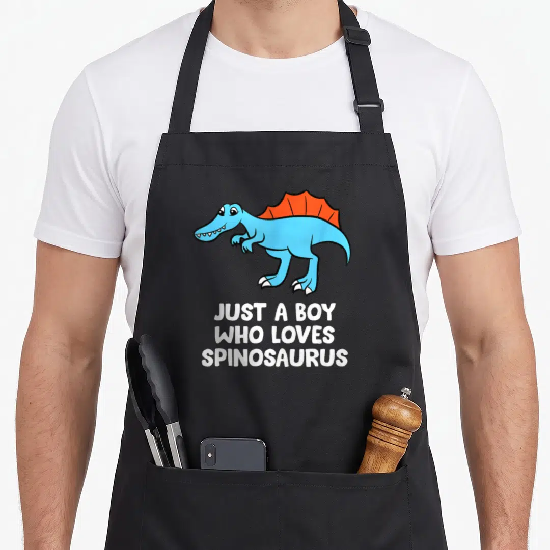 Retro spinosaurus dinosaur apron - Blue cartoon Spinosaurus with orange sail and white text JUST A BOY WHO LOVES — shown on a black cotton canvas apron against a white background
