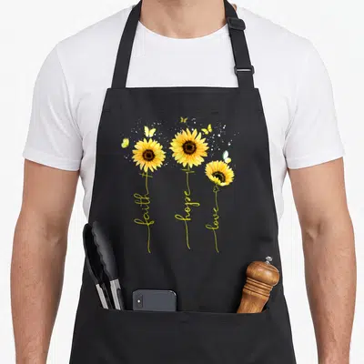 Faith sunflower apron - Three yellow sunflowers with stems spelling faith hope love and scattered butterflies — shown on a black cotton canvas apron against a white background