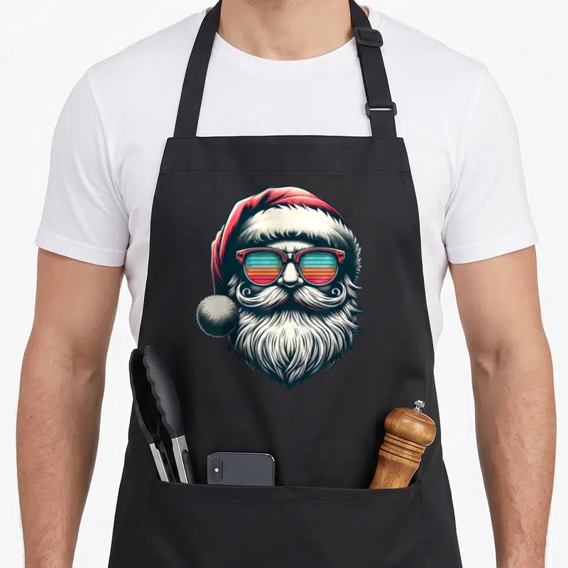 Sunglass christmas santa apron - Cartoon Santa with red hat, striped sunglasses, white beard, no text — shown on a black cotton canvas apron against a white background