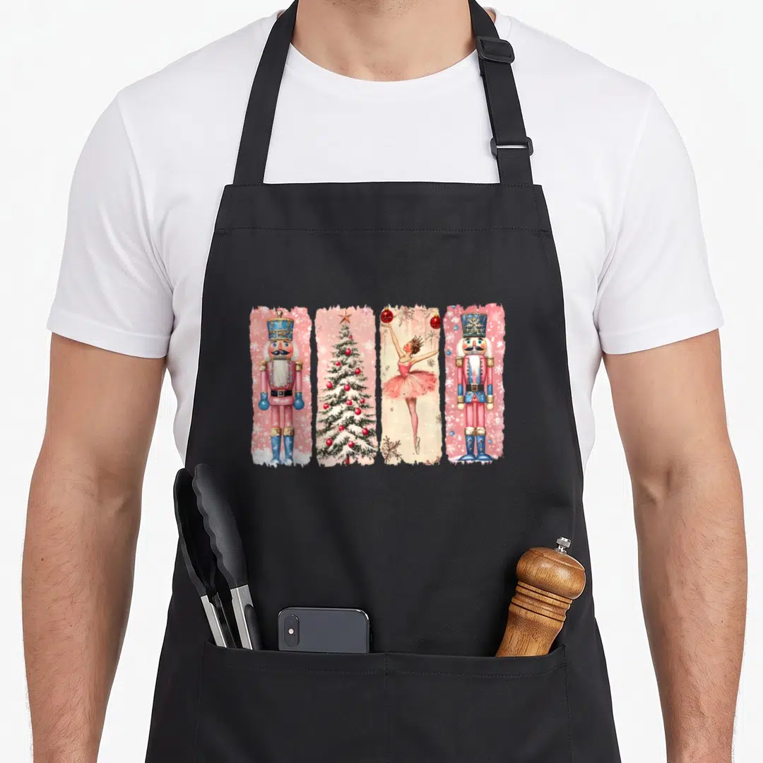 Nutcracker pink christmas apron - Four pink panels showing nutcracker soldiers, a Christmas tree, and a ballerina — shown on a black cotton canvas apron against a white background