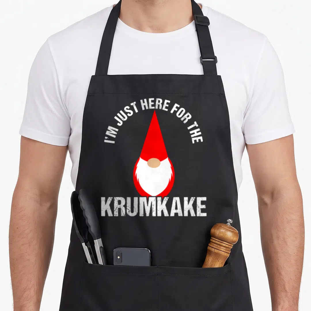 Krumkake scandinavian aprons - Cartoon gnome with red hat and white text reading I'M JUST HERE FOR THE KRUMKAKE — shown on a black cotton canvas apron against a white background