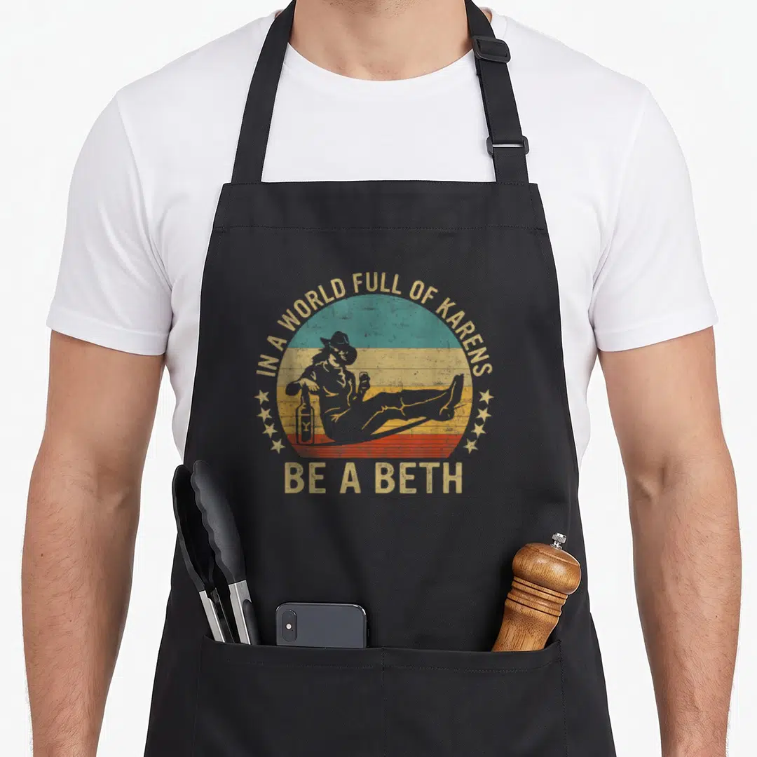 Karen beth novelty apron - Retro sunset graphic with woman silhouette and text In A World Full Of Karens Be A Beth — shown on a black cotton canvas apron against a white background