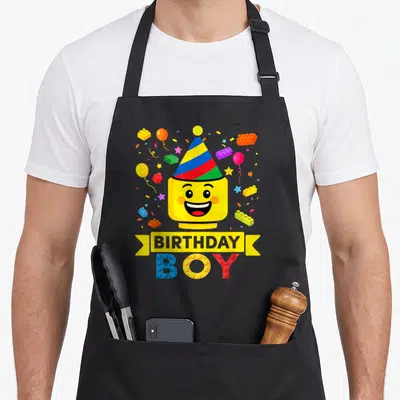 Hat building aprons - Smiling yellow toy figure in striped party hat with BIRTHDAY BOY text and scattered building — shown on a black cotton canvas apron against a white background