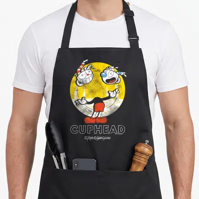 Cuphead juggling apron - Two cartoon characters in yellow circle with red shorts and stacked CUPHEAD text below — shown on a black cotton canvas apron against a white background