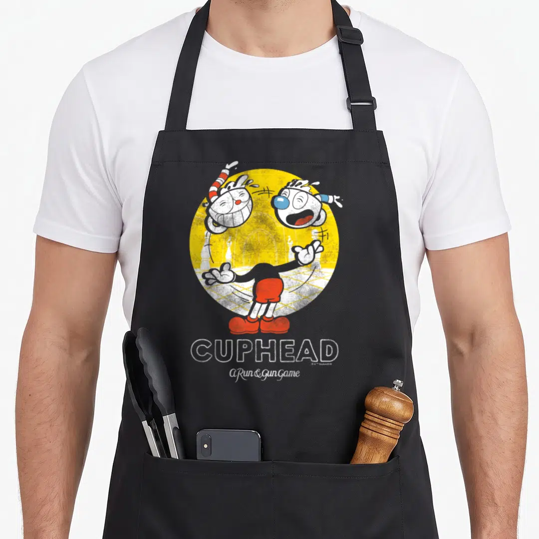 Cuphead juggling apron - Two cartoon characters in yellow circle with red shorts and stacked CUPHEAD text below — shown on a black cotton canvas apron against a white background