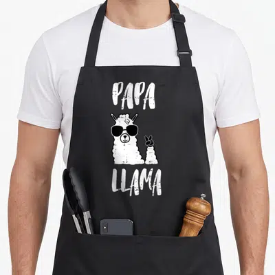 Llama father day apron - PAPA LLAMA text above a white cartoon llama wearing black sunglasses and holding up a hoof — shown on a black cotton canvas apron against a white background