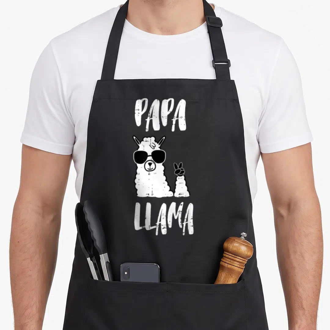Llama father day apron - PAPA LLAMA text above a white cartoon llama wearing black sunglasses and holding up a hoof — shown on a black cotton canvas apron against a white background
