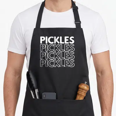 Cute pickle apron - Stacked white text reading PICKLES four times, top line solid, three below outlined — shown on a black cotton canvas apron against a white background