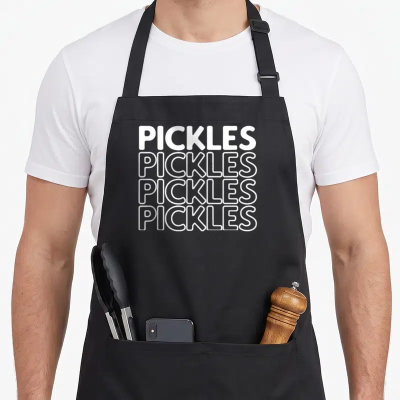 Cute pickle apron - Stacked white text reading PICKLES four times, top line solid, three below outlined — shown on a black cotton canvas apron against a white background
