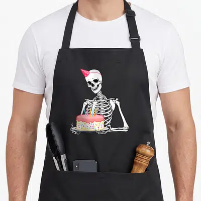 Birthday outfit funny halloween apron - Cartoon skeleton in pink hat holding cake with sprinkles and candles — shown on a black cotton canvas apron against a white background