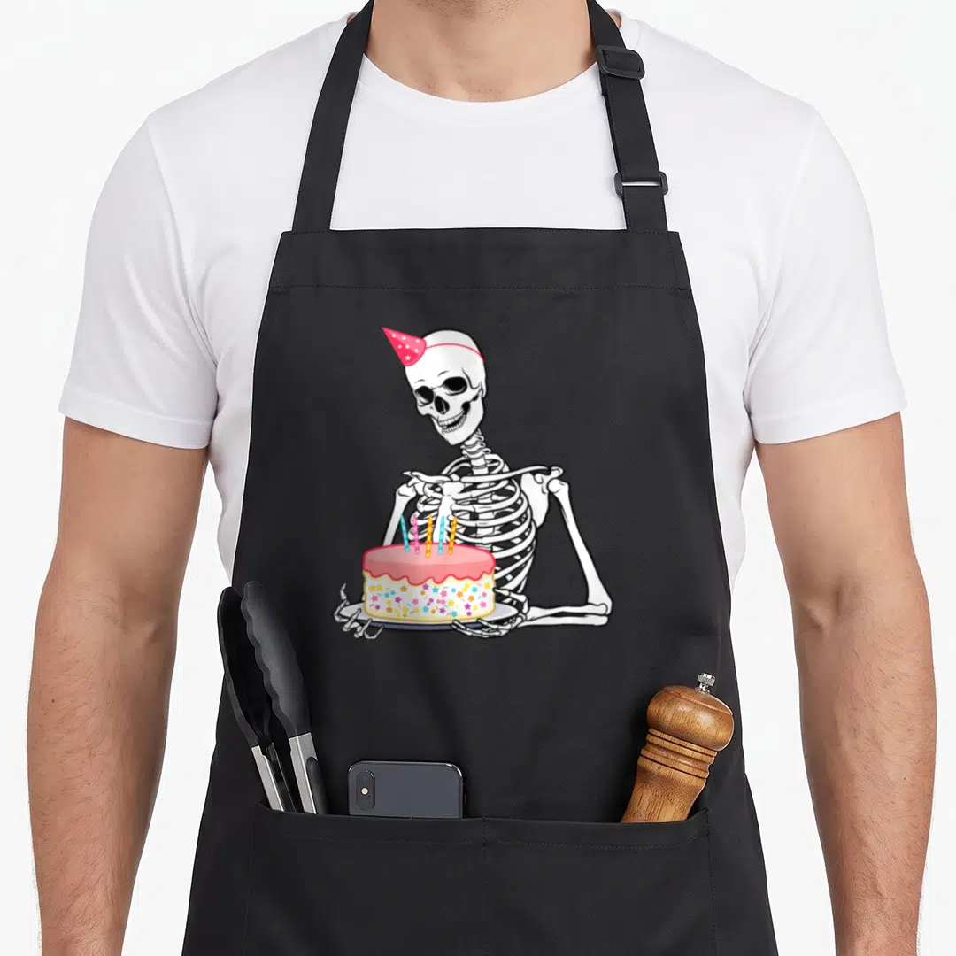 Birthday outfit funny halloween apron - Cartoon skeleton in pink hat holding cake with sprinkles and candles — shown on a black cotton canvas apron against a white background