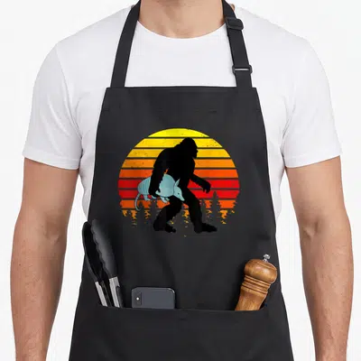 Cradling bigfoot apron - Black bigfoot silhouette holding light blue armadillo against yellow orange red striped sun — shown on a black cotton canvas apron against a white background
