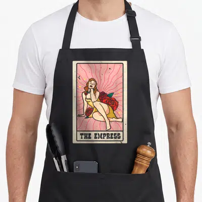 Empress shaman apron - Retro illustration of a woman in a yellow dress lying among red roses against a pink background — shown on a black cotton canvas apron against a white background