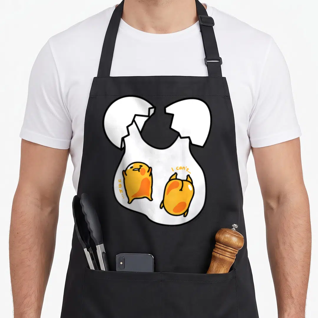 Twin egg apron - Two yellow egg characters in cracked shell with ugh... and I can't... text — shown on a black cotton canvas apron against a white background